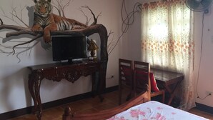A bedroom with a bed, a TV on a wooden stand, a chair, a table, a fan, and a tiger mural on the wall.