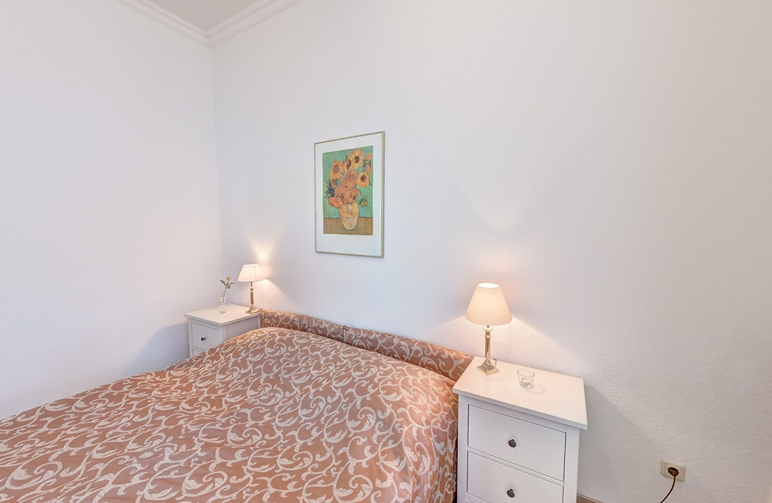 Ein Schlafzimmer mit einem gemusterten Bettbezug, zwei Nachttischen mit Lampen, einem Wandbild und einer Steckdose.