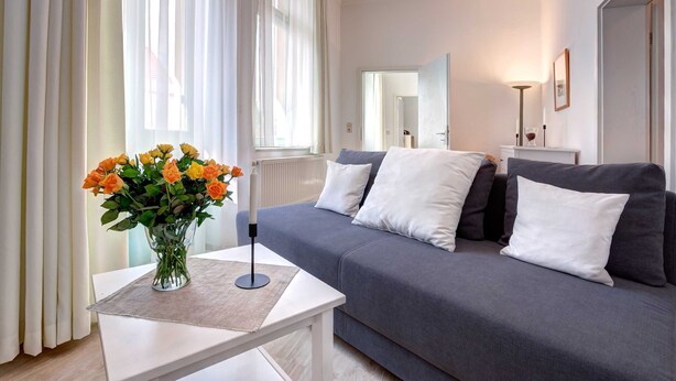A modern living room with a grey sofa, white cushions, a small table with a vase of flowers, and a large window with sheer curtains. - Villa Ostseewarte Whg. 6, TERRASSE - Villa Ostseewarte, STRANDNAH, teilw. (Heringsdorf)
