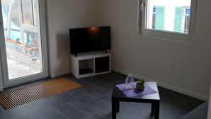 Ein modernes Wohnzimmer mit einem Flachbildfernseher auf einem weißen Schrank, einem kleinen Tisch mit einer Pflanze und einem großen Fenster mit Blick ins Freie.