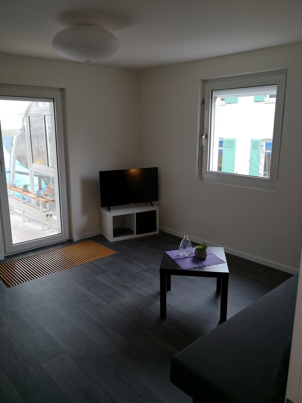 A modern living room with a flat-screen TV on a white stand, a small table with a plant, and a large window offering a view of the outside. - very central and very quiet, apartment in the heart of Lindau (Lindau (Bodensee))