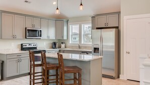 A modern kitchen with grey cabinets, a stainless steel refrigerator, a microwave, and a large island with bar stools. - Good To Be Back: Soundside, Private Pool & Spa, Great Community Amenities (Corolla)