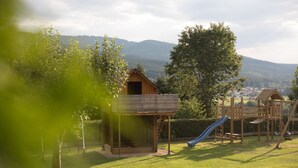 A wooden playhouse with a slide, set in a grassy area with trees and mountains in the background. - Appartementhaus Weber (Arrach)