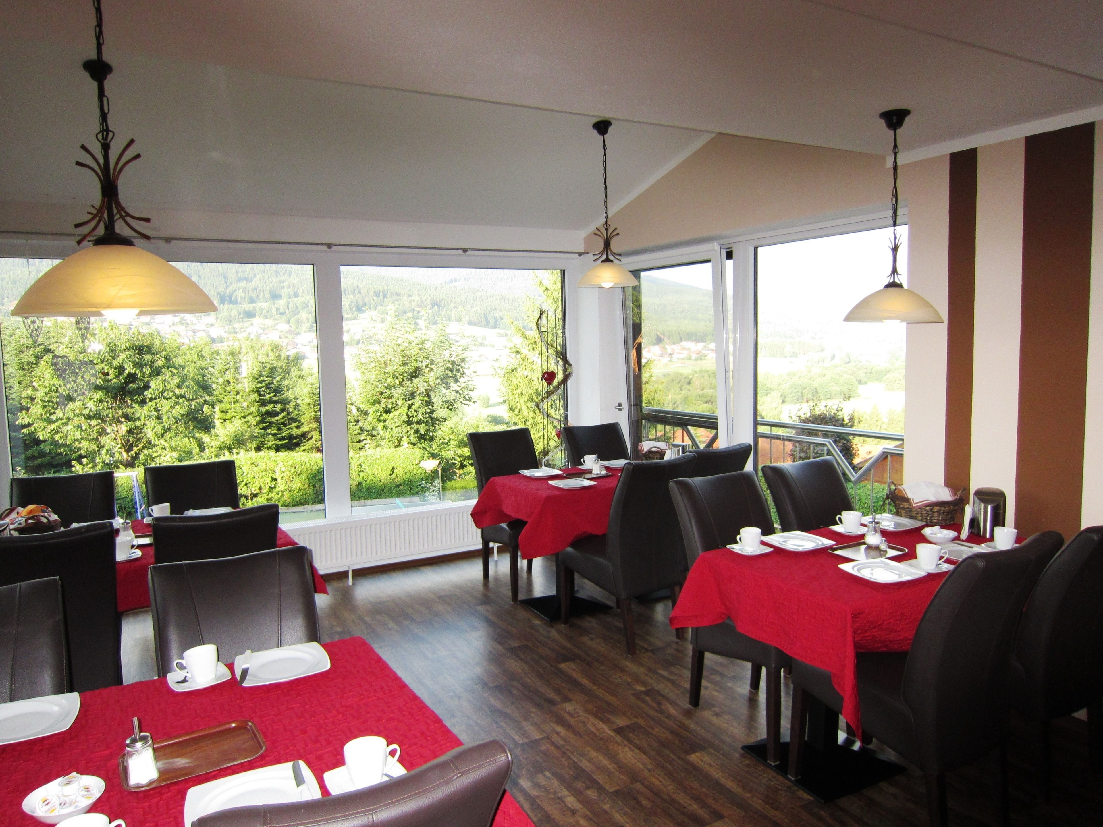 A restaurant with tables set for dining, large windows offering a view of greenery, and hanging pendant lights.