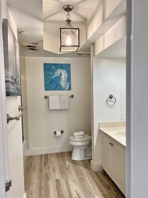 Ein Badezimmer mit Holzboden, Toilette in Weiß, Waschbecken mit Spiegel, Handtuchhalter und einer Deckenleuchte.