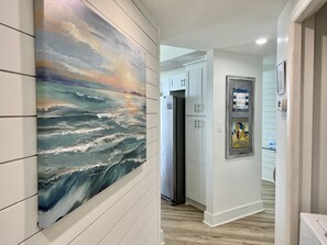 Um corredor com uma grande pintura abstrata, uma geladeira, uma porta com uma placa de boas-vindas e piso de madeira.