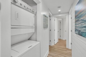 Un cuarto de lavandería con lavadoras y secadoras apiladas, un pasillo con puertas y un cuadro en la pared.