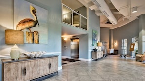 A spacious lobby with a high ceiling, a wooden console table with a lamp, a painting of a pelican, and an elevator in the background.