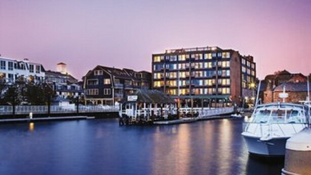 A waterfront hotel with multiple floors, a dock with boats, and a calm water body reflecting the buildings.