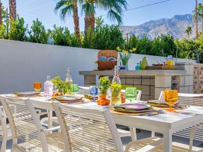 Un espacio de comedor al aire libre con una mesa preparada para cuatro personas, rodeada de sillas y una parrilla de barbacoa al fondo.