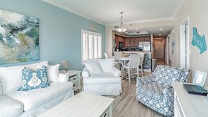 Un amplio salón con un sofá blanco, un sillón estampado y una mesa de comedor. Se ve una cocina al fondo con armarios de madera y una encimera.