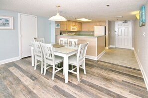 A dining area with a glass table and white chairs, a kitchen with wooden cabinets and a white refrigerator, and a hallway with a wooden floor. - Oceanfront Views from Every Room in This Condo (Myrtle Beach)