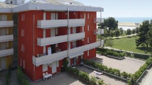 A red and white multi-story building with balconies, a paved courtyard, and a view of the sea. - Studio in a seafront building-Beahost (Bibione)