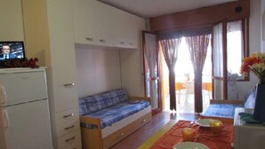 A compact room with a bed, a small table with a red tablecloth, and a kitchenette area. - Studio in a seafront building-Beahost (Bibione)