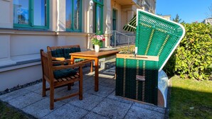 Et sæt af træbænk og bord på en belagt terrasse med en grøn stol foran en bygning med grønne vinduesrammer.