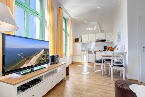 A modern living room with a flat-screen TV on a wooden console, a dining area with white chairs, a kitchen with white cabinets, and large windows with curtains.