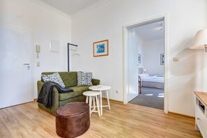 A room with a green sofa, a small round table, a brown ottoman, a white lamp, and a door leading to another room with a bed.