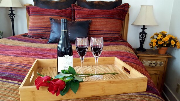 Una bandeja de madera con una botella de vino, dos copas de vino tinto y un ramo de rosas rojas sobre una cama con ropa de cama a rayas y almohadas.