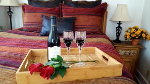 Un plateau en bois avec une bouteille de vin, deux verres de vin rouge et un bouquet de roses rouges, le tout posé sur un lit à la literie et aux oreillers à rayures.