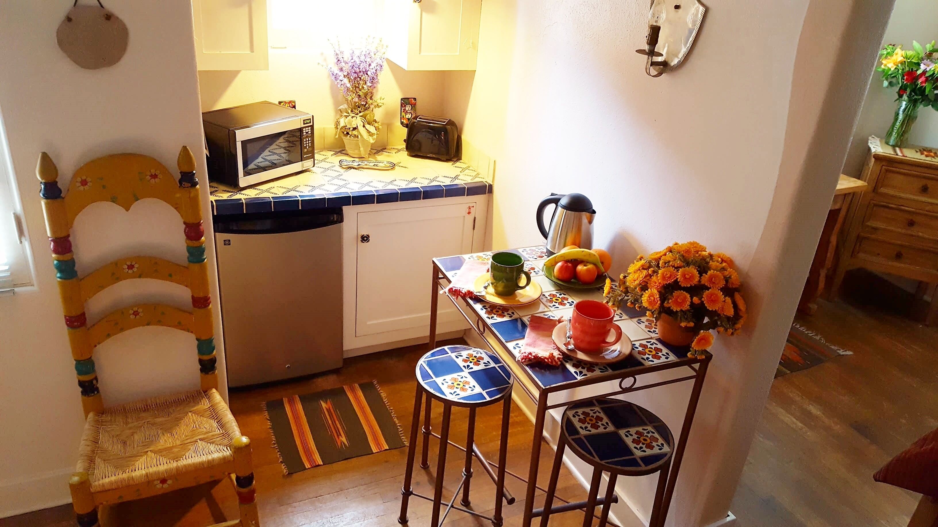 A compact kitchen with a microwave, toaster, and a small table with a coffee pot, fruit bowl, and flowers.