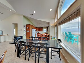 A spacious dining area with a large table and chairs, a kitchen in the background, and a view of the beach through large windows.