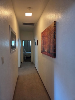 Un couloir avec un luminaire au plafond, une grille d’aération, des tableaux encadrés et une peinture au mur.