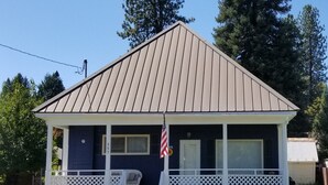 A small, single-story house with a dark blue exterior, a white porch with lattice railings, and a brown roof. - The Mountain House at the base of Mount Shasta (McCloud)