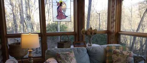 A cozy room with a large window, a wooden table with a lamp, a cushioned sofa, and a vase with dried flowers.