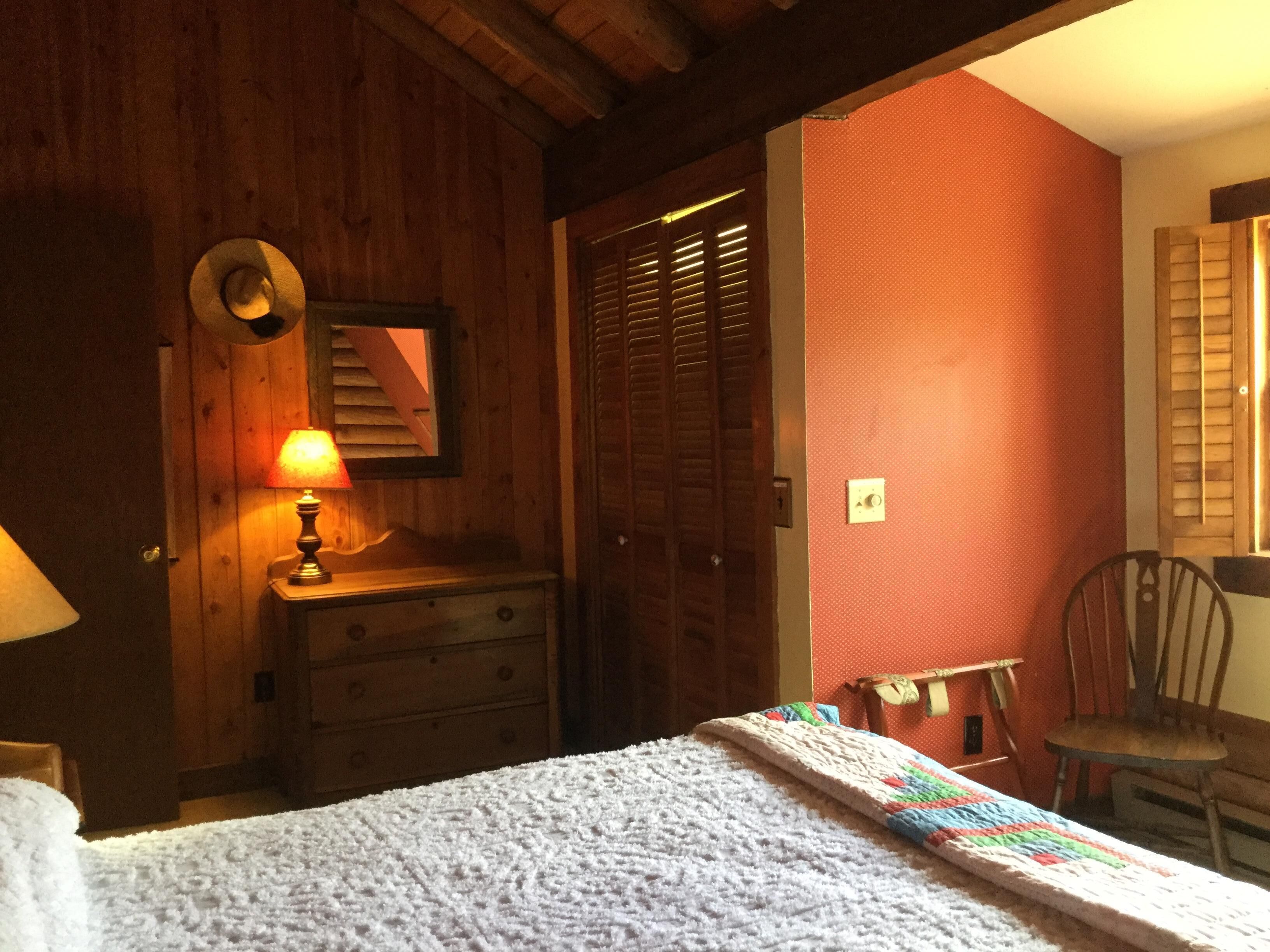 A bedroom with wooden walls, a bed with a white blanket, a wooden dresser, a lamp, and a chair.