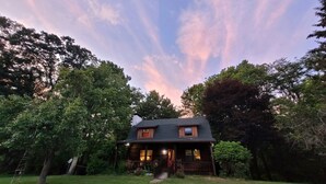 Une cabane en rondins avec un porche, entourée d’arbres et bordée d’une pelouse.