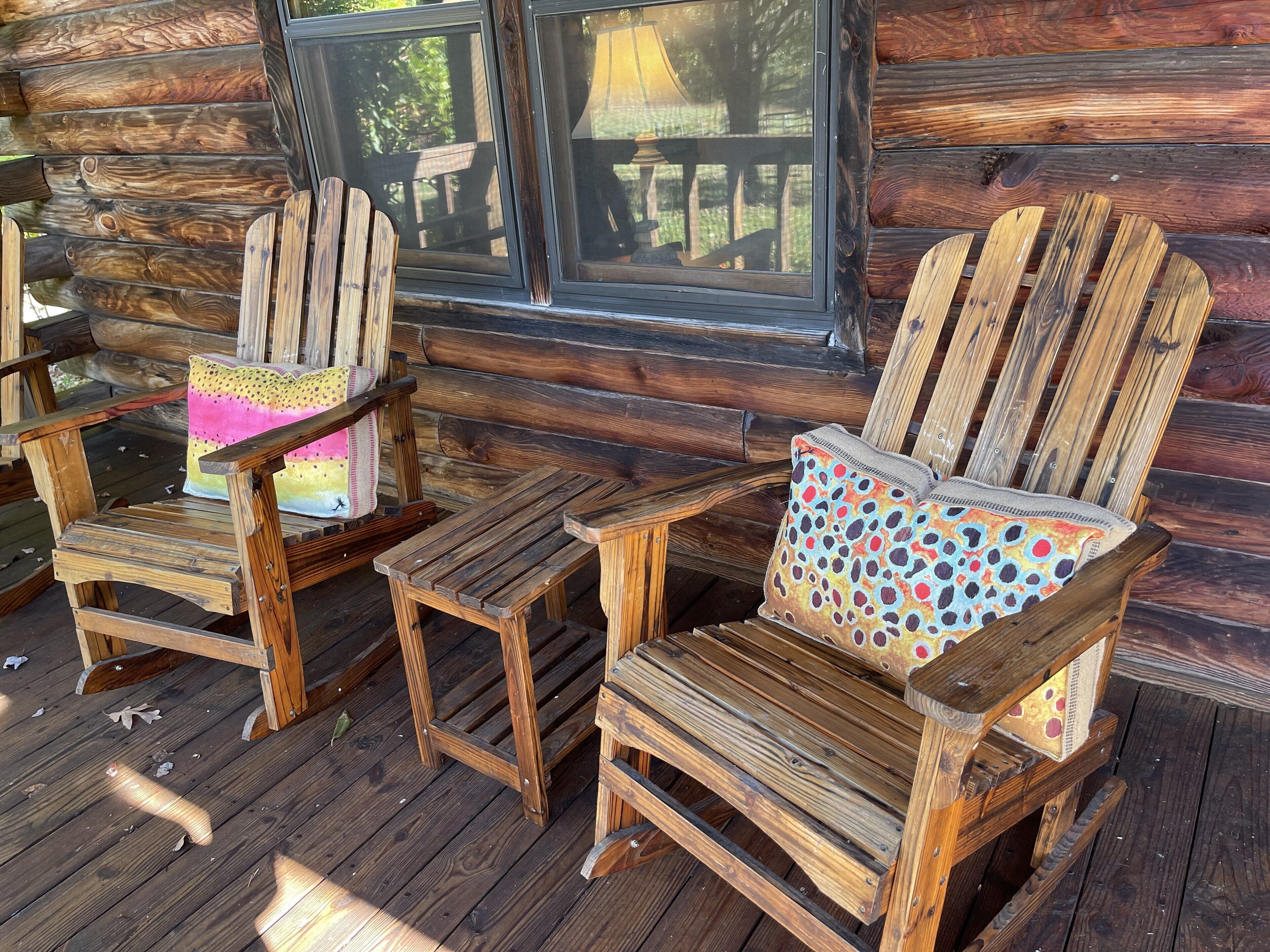 Two Adirondack chairs with colorful cushions on a wooden deck in front of a log cabin with a window.
