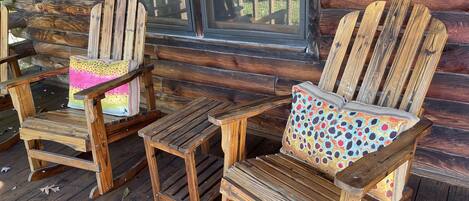 Two Adirondack chairs with colorful cushions on a wooden deck in front of a log cabin with a window.