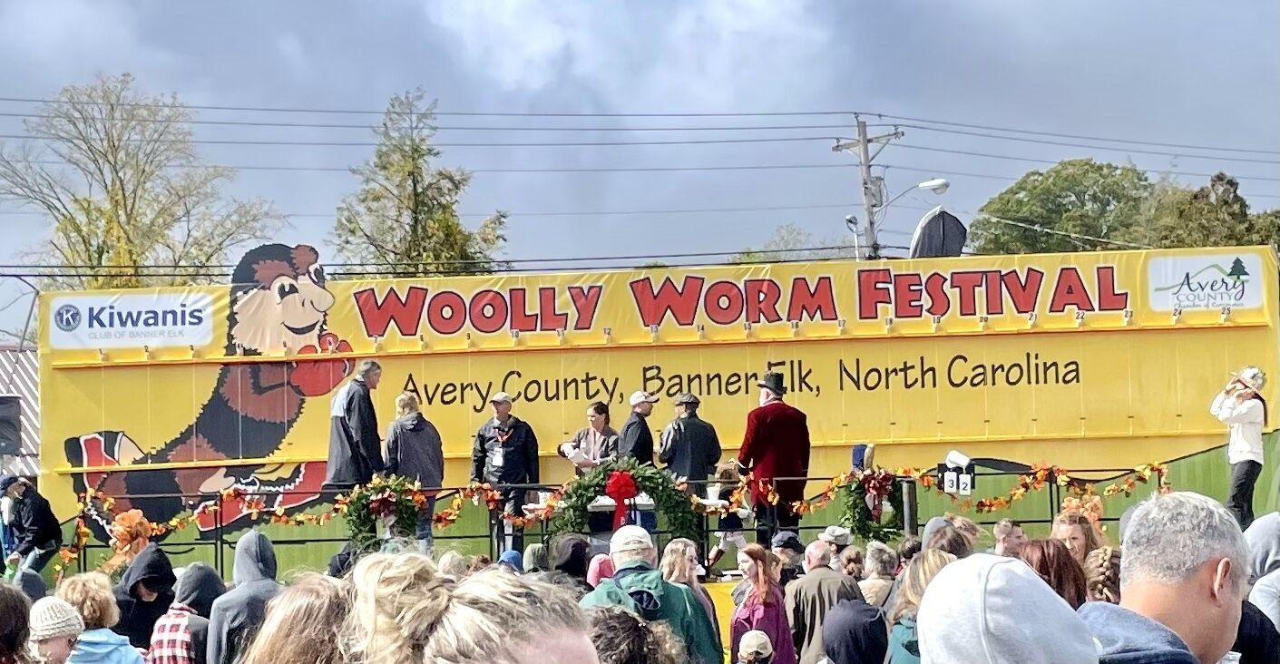 A large yellow banner with 'WOOLLY WORM FESTIVAL' text, featuring a cartoonish worm character and 'Avery County, Banner Elk, North Carolina' text.