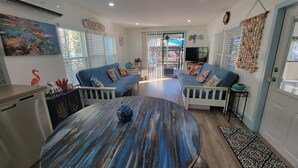 Amplia sala de estar con una mesa redonda de madera, dos sofás azules con barandillas blancas, una zona de cocina con refrigerador y un ventanal con persianas.