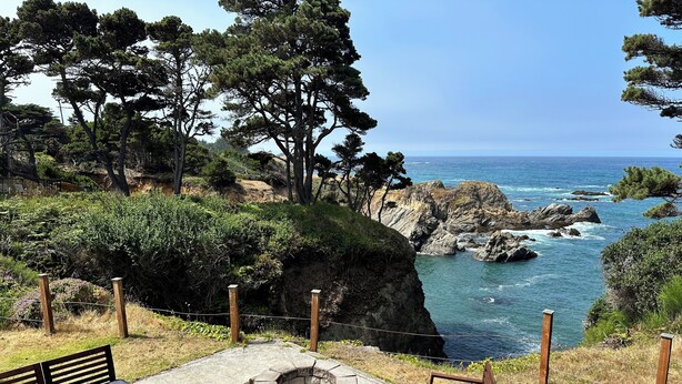 A patio with outdoor furniture, a fire pit, and a view of the ocean and rocky coastline. - Secluded Ocean View Mid-Century Modern Retreat  (Fort Bragg)