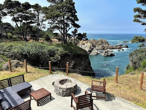 A patio with outdoor furniture, a fire pit, and a view of the ocean and rocky coastline. - Secluded Ocean View Mid-Century Modern Retreat  (Fort Bragg)