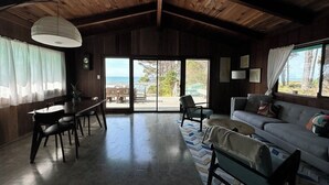 A spacious room with a wooden ceiling, a dining area with a table and chairs, a sofa, and a view of the outdoors through large windows. - Secluded Ocean View Mid-Century Modern Retreat  (Fort Bragg)