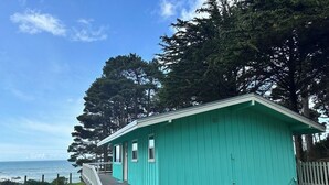 A turquoise building with a wooden deck, a white fence, and a view of the ocean. - Secluded Ocean View Mid-Century Modern Retreat  (Fort Bragg)