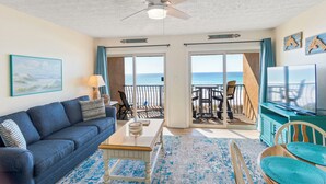 A living room with a blue sofa, a coffee table, and a television. There is a balcony with a view of the ocean. - 26- Your vacation starts here in this FAMILY FRIENDLY condo that sleeps 6! (Destin)