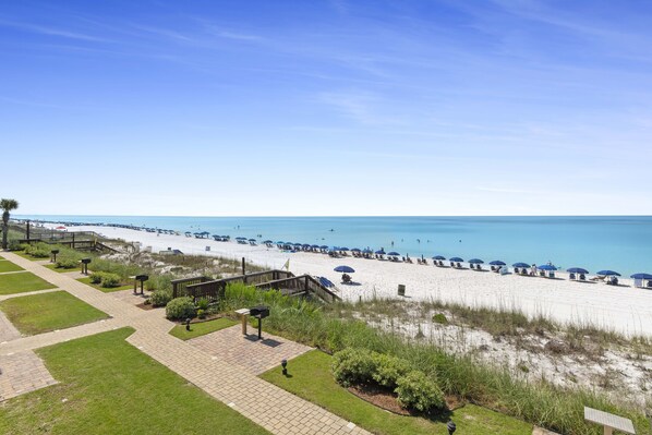 A beach view with a pathway leading to a beach area, featuring umbrellas and beach chairs.