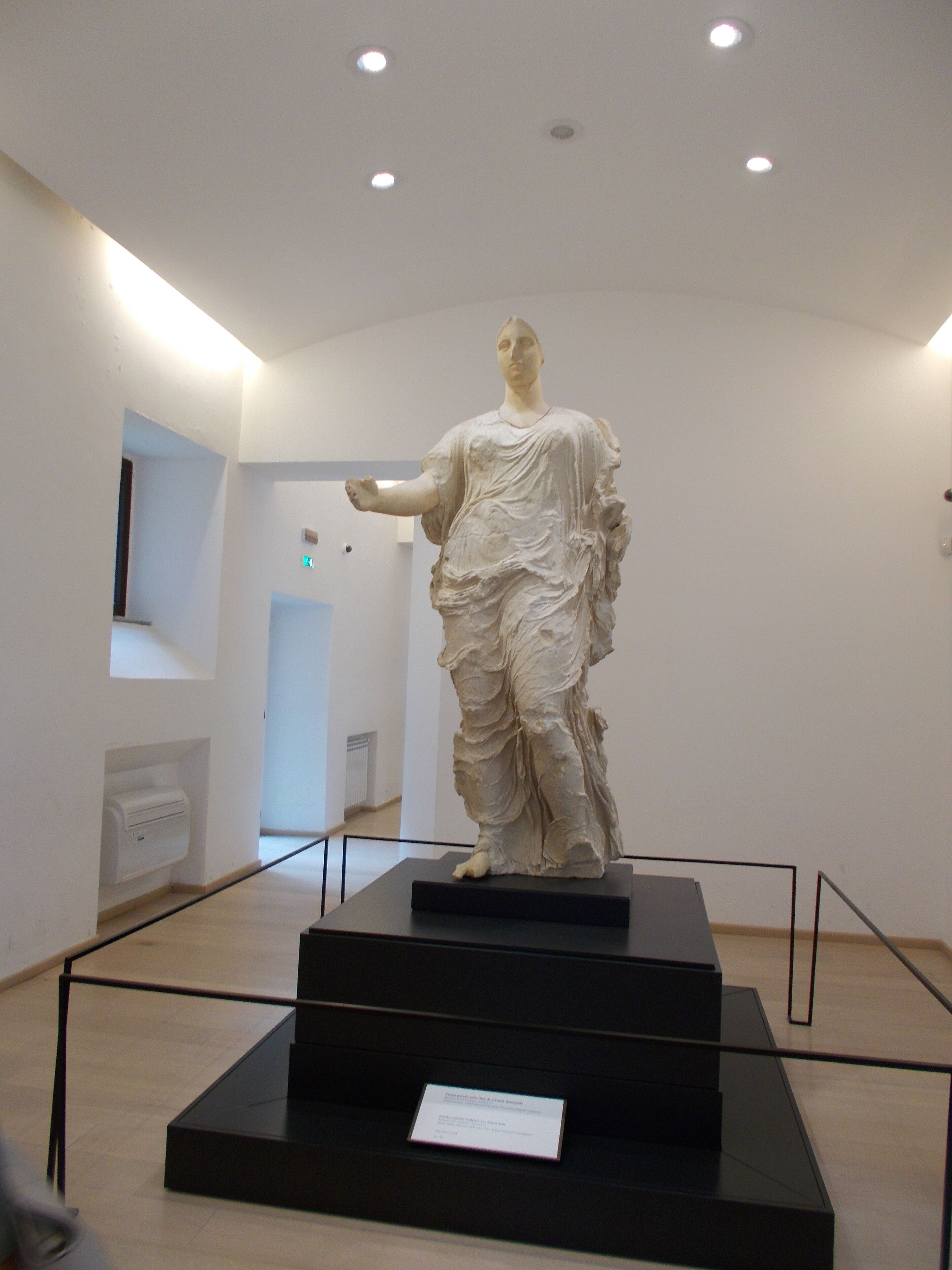 A white marble statue on a black pedestal in a museum with white walls and a high ceiling.