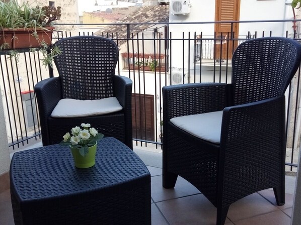 Two wicker chairs with cushions on a balcony with a tiled floor and a small table with a flower pot.
