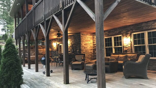 Une terrasse extérieure couverte, dotée d’un plancher en bois, de murs en pierre et d’un espace salon avec des meubles en osier.
