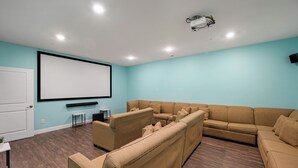 Una sala home theater con un grande schermo, un divano a forma di L color marrone e un tavolino con diffusori.