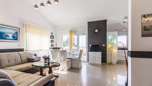 Ett modernt vardagsrum med en soffa, ett matbord och en tv. Det finns en balkong med utsikt över havet.