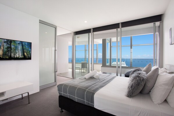 A modern bedroom with a large bed, a flat-screen TV, and a balcony with a view of the sea.
