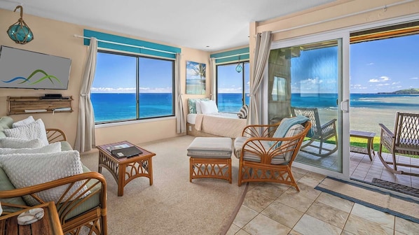 A room with a bed, a sofa, wicker chairs, a small table, and large windows with a view of the ocean.