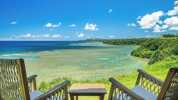 A deck with chairs overlooking a clear blue sea and a lush green hillside.