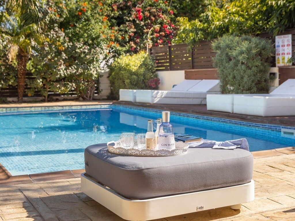 A poolside area with a cushioned seating area, a tray with bottles and glasses, and a wooden fence with greenery in the background.