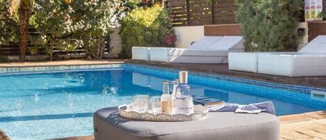 A poolside area with a cushioned seating area, a tray with bottles and glasses, and a wooden fence with greenery in the background.